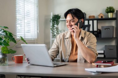 Tatmin olmuş, mutlu bir Asyalı işadamı, müşterisiyle telefonda konuşurken bilgisayarını tamir ediyor, ofisinde telefonla iş yapıyor..