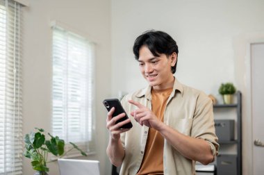 Günlük giysiler içinde pozitif Asyalı bir işadamı ofisinde duruyor ve telefonundaki mesajları ya da sohbetleri okurken gülümsüyor. İnsanlar, kablosuz teknoloji.