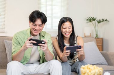 Neşeli, mutlu bir Asyalı çift telefonlarıyla oyun oynamaktan, birlikte kanepede oturmaktan ve evde eğlenmekten zevk alır..
