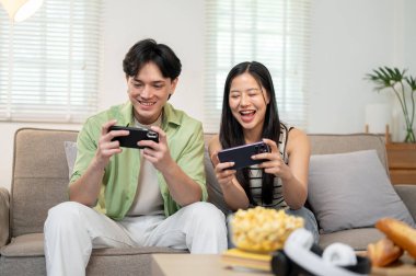 Neşeli, mutlu bir Asyalı çift telefonlarıyla oyun oynamaktan, birlikte kanepede oturmaktan ve evde eğlenmekten zevk alır..