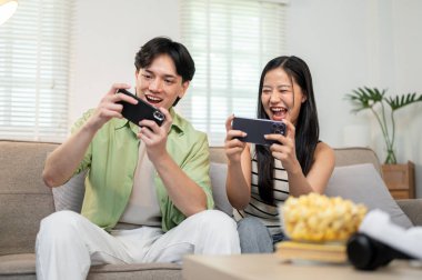 Neşeli, mutlu bir Asyalı çift telefonlarıyla oyun oynamaktan, birlikte kanepede oturmaktan ve evde eğlenmekten zevk alır..