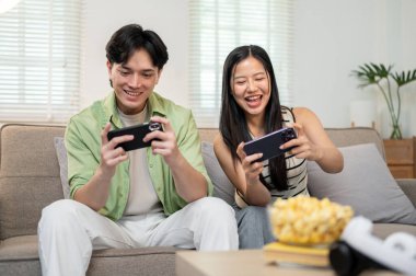 Neşeli, mutlu bir Asyalı çift telefonlarıyla oyun oynamaktan, birlikte kanepede oturmaktan ve evde eğlenmekten zevk alır..