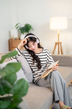 Güzel ve düşünceli, rahat giysiler içinde, kulaklık takan genç bir Asyalı kadın minimalist evindeki kanepede günlüğünü yazarken derin düşüncelere dalıyor..
