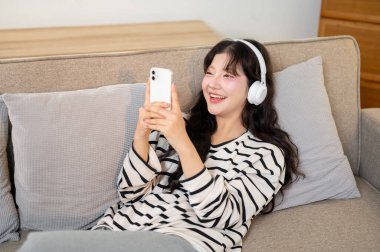 Çekici genç bir Asyalı kadın oturma odasında bir kanepede dinleniyor, akıllı telefonuyla arkadaşlarıyla sohbet ederken kulaklıklarıyla müziğin tadını çıkarıyor..