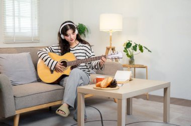 Güzel, rahat giyinmiş, kulaklık takan Asyalı genç bir kadın oturma odasındaki bir kanepede otururken gitar çalmaya ya da pratik yapmaya odaklanmış..