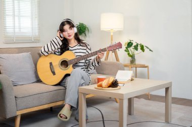 Güzel, rahat ve rahat bir Asyalı kadın dairesindeki bir kanepede gitar çalarken kulaklıkla müzik dinliyor..