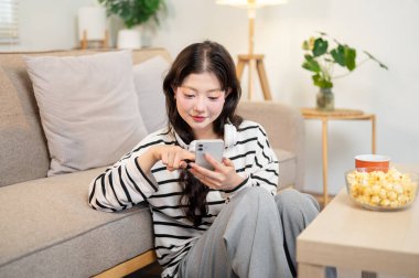 Çekici, sakin Asyalı bir kadın sabah oturma odasında dinlenirken sohbet okuyor ya da akıllı telefonuyla kayıyor. İnsanlar, yaşam tarzları, kablosuz teknoloji, ev hayatı