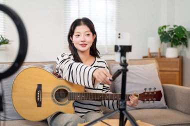 Kendine güvenen, neşeli Asyalı bir kadın nüfuzlu kişi, takipçileriyle iletişime geçmek için sosyal medyada canlı yayın yaparken, oturma odasında gitar çalıyor..