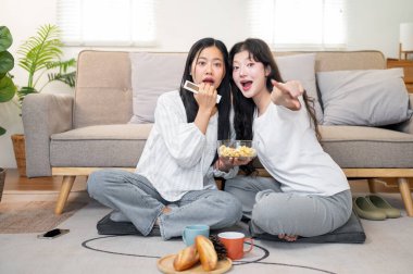 İki neşeli ve neşeli genç Asyalı kadın evlerinin rahat oturma odasında film izlerken mutlu bir şekilde patlamış mısırın tadını çıkarıyorlar..