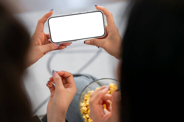 Крупным планом две подруги разговаривают во время просмотра фильма на смартфоне на диване в гостиной. смартфон с белым макетом экрана