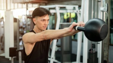 Spor giyim sektöründe kararlı ve güçlü bir Asyalı adam spor salonunda spor malzemeleriyle egzersiz yaparken güçlü kol kaslarıyla kettlebell ile çalışıyor. ağırlık antrenmanı konsepti