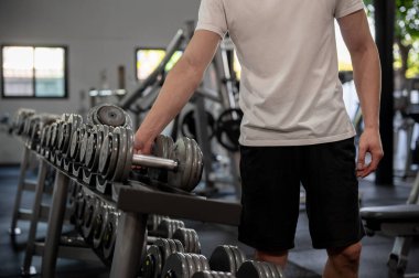 Spor giyimli güçlü ve sportif Asyalı bir adam spor salonundaki salondan ağırlık antrenmanına hazırlanan bir dambıl seçiyor. yaşam tarzları, ağırlık eğitimi, fitness ekipmanları