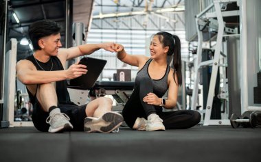 Neşeli bir Asyalı erkek fitness eğitmeni ve bayan müşterisi spor salonunda yerde oturmuş antrenmanlarını bitirdikten sonra birbirlerini yumrukluyorlar. Fitness hayatı, sağlıklı yaşam tarzı