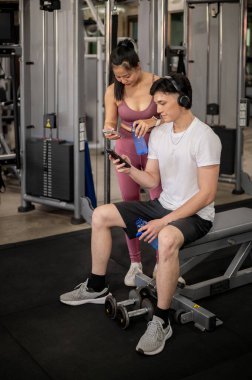 Pozitif Asyalı bir erkek arkadaş ve kız arkadaş, spor kıyafetleri içinde spor salonunda, spor yaptıktan sonra dinleniyorlar, akıllı telefonlarındaki mesajları dinliyorlar..