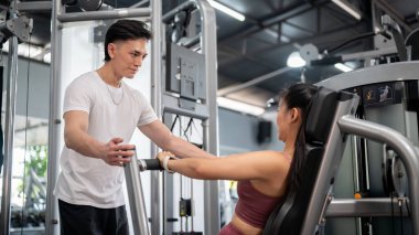 Destekleyici ve profesyonel bir Asyalı erkek fitness eğitmeni, bayan bir müşteriye göğüs pres makinesini kullanma ve spor salonunda formda kalma talimatı veriyor..
