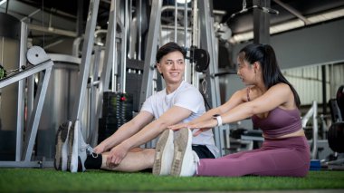 Spor kıyafetli mutlu bir Asyalı kız arkadaşıyla spor salonunda ısınırken sohbet ediyor. İkisi de antrenmana başlamadan önce bacaklarını esnetiyorlar..