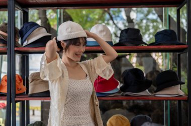 Güzel ve tatmin olmuş Asyalı bir kadın şapka dükkanında şapka deniyor, almak için eşyalar seçiyor ve hafta sonları alışveriş çılgınlığının tadını çıkarıyor. Yaşam tarzı, tatil, moda