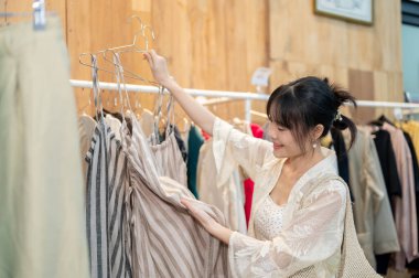 Mutlu bir Asyalı bayan müşteri bir giyim mağazasından almak için eşyalar seçiyor, tek başına alışveriş kıyafetlerinin keyfini çıkarıyor. Yaşam tarzı, insanlar, tüketim