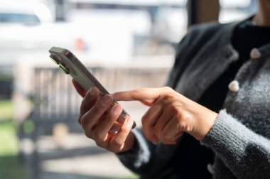 Kapalı alanda otururken akıllı telefonunu kullanan, parmaklarını ekranda gezdiren bir kadının yakın çekimi. İnsanlar ve kablosuz teknoloji kavramları