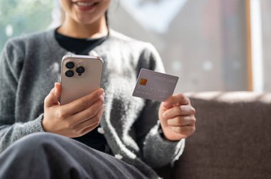 Gündelik kıyafetlerle çekilmiş bir kadın aydınlık bir günde elinde akıllı telefon ve kredi kartıyla evde oturur. Çevrimiçi alışveriş, çevrimiçi ödeme, mobil bankacılık, nakitsiz, finansal