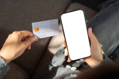 Evin içinde oturan bir kadının üst görüntüsü, elinde kredi kartı ve elinde beyaz ekran maketi olan bir akıllı telefon. Çevrimiçi alışveriş, çevrimiçi ödeme, mobil bankacılık, nakitsiz, finansal