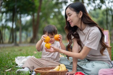 Güzel ve ilgili bir Asyalı anne, küçük oğluyla halka açık bir parkta piknik yaparken meyve kullanarak oynar. Annelik, çocukluk, anaokulu, mutlu aile.
