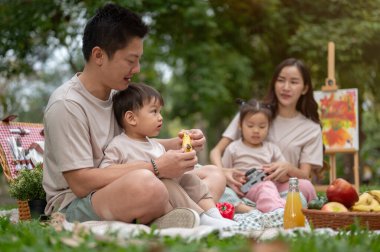 Şefkatli bir Asyalı baba, yeşil bir parktaki aile pikniğinde kucağında küçük oğluyla onun için muz soyarken. Aile, babalık, çocukluk