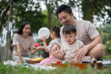 İlgili bir Asyalı baba, anne ve kızı arka planda atıştırmalık hazırlarken, parkta ailesiyle piknik yaparken küçük oğluna tuvale resim çizmeyi ve resim yapmayı öğretir..