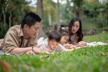 İlgili bir Asyalı baba küçük oğluna kağıt çizmeyi öğretirken ikisi de piknik battaniyesine uzanmış, ailesiyle parkta piknik yaparken eğleniyor..