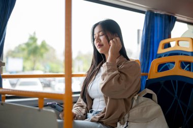 Rahat giyinmiş Asyalı bir kadın, şehirde işe gidip gelirken bir otobüsün camında oturup kulaklıklarıyla müzik dinlemekten hoşlanır. şehir hayatı, toplu taşıma