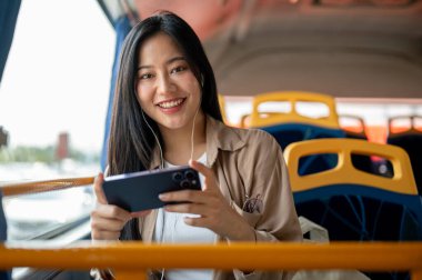Neşeli, güzel, sıradan kıyafetler içinde Asyalı bir kadın bir otobüsün cam kenarında otururken akıllı telefonuyla film ya da video izlemekten zevk alıyor..