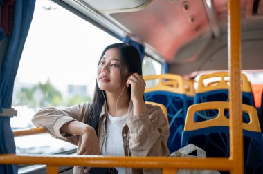 Güzel, rahat bir Asyalı kadın, sıradan kıyafetler içinde bir otobüsün camında otururken kulaklıklarıyla müzik dinliyor. şehir hayatı, toplu taşıma