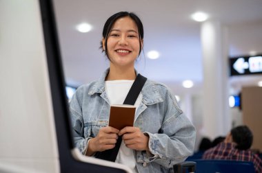 Kot ceketli güzel bir Asyalı kadın havaalanındaki bir gişede kameraya gülümseyip bir yerlere seyahat ederken pasaportunu tutuyor. seyahat, tatil, ulaşım