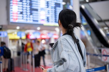 Asyalı bir bayan yolcunun havaalanındaki uçuş bilgilerine baktığı, uçuş zamanını kontrol ettiği, seyahat etmeye hazır olduğu görüntüsü..