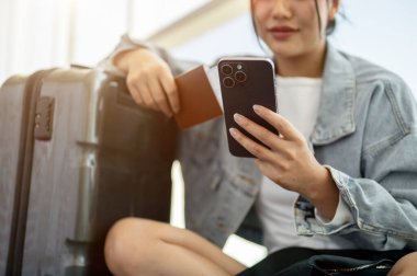 Pozitif bir Asyalı kadın yolcu havaalanı terminalinde bavulunun yanında oturmuş, uçuş zamanını beklerken akıllı telefonuyla sohbet ediyor..