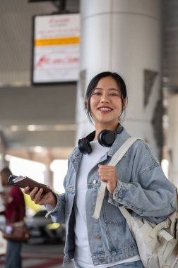 Kot ceketli ve sırt çantalı çekici, gülümseyen Asyalı bir bayan yolcu havaalanının buluşma noktasında duruyor ve gideceği yere bir taksi gelmesini bekliyor. insanlar ve ulaşım