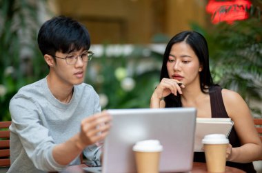Çekici bir Asyalı kadın meslektaşının fikirlerini dikkatle dinliyor, bir kahve dükkanındaki açık hava masasında çalışırken bilgisayarın ekranına bakıyor, uzaktan çalışıyor..