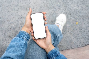 Akıllı telefonu tutan bir çift elin en üst görüntüsü. Kot giyin ve tahta bankta bağdaş kurarak otur..