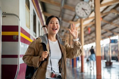 Gülümseyen Asyalı kadın, mutlu ve heyecanlı görünüyor, tren istasyonunda arabadan indikten sonra elini sallıyor. Klasik tren istasyonu ya da tren istasyonu.