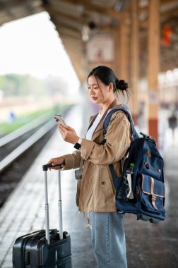 Asyalı kadın tren istasyonunda ya da tren istasyonunda çanta ve bavulla durduğu sırada telefona bakıyor..
