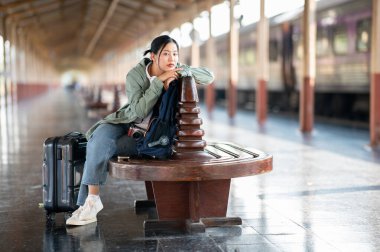 Asyalı bir kadın, tren istasyonunda ya da tren istasyonunda bavulu ve çantasıyla tahta bankta bacak bacak bacak üstüne atmış otururken sıkılmış görünüyor..