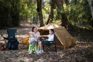 Anne ve oğlu kamp çadırının önünde el ele tutuşurken konuşuyorlar. Aile gezisi, orman kampı, açık hava aktiviteleri..
