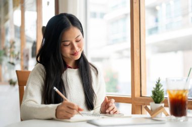 Asyalı bir kadın deftere yazarken gülümsüyor, büyük pencere duvarlarının yanındaki masada oturuyor. Kafede ders çalışmak ya da ödev yapmak.