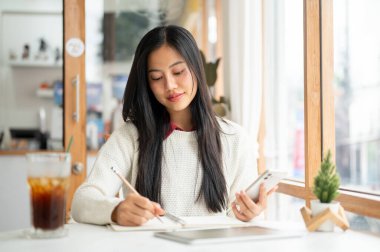 Asyalı bir kadın elinde bir tablet ve masada bir içki yeriyle telefonu tutarken deftere bir şeyler yazıyor. Telefondan not almak ya da kafeden birini aramak..