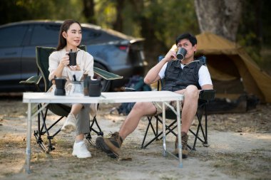 Bir adam, kadının kamp çadırlarının önündeki katlama masasında onun için kahve koyduğu bardaktan içiyor. Birlikte seyahat, orman kampı, açık hava aktivitesi..