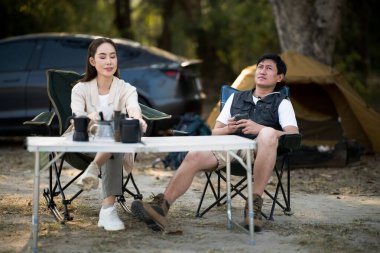 Bir adam ve bir kadın, kamp çadırlarının önündeki katlama masasında birlikte oturuyorlar. Birlikte seyahat, orman kampı, açık hava aktivitesi..