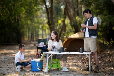 Baba ve anne masayı hazırlarken oğullarına gülümsüyor, çaydanlık, kahve makinesi ve kupalarla. Aile gezisi, orman kampı, açık hava aktiviteleri..