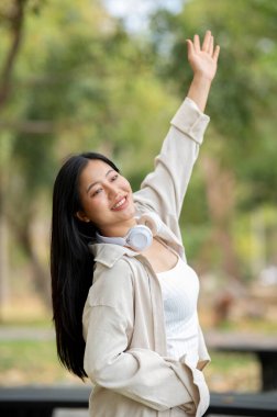 Asyalı bir kadın tek kolu havada duruyor ve parkta ileriye bakıyor, temiz hava soluyor. Doğada dinlenme, açık hava aktivitesi, yeşil orman parkı..