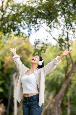 Gülümseyen Asyalı bir kadın iki kolu havada parkta temiz hava soluyor. Doğada dinlenme, açık hava aktivitesi, yeşil orman parkı..