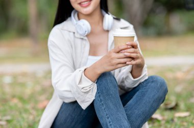 Gülümseyen Asyalı bir kadının iki eliyle fincanı tutarken çim tarlasında dizlerinin üzerinde oturmasını yakından çek. Doğada dinlenme, açık hava aktivitesi, yeşil orman parkı..